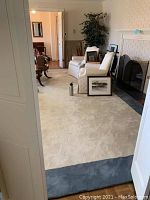 A wide view of the large rectangular cream area rug laid out in a living room with white seating furniture and fireplace, showing overall size, shape, and color with grey-blue border visible on one end.