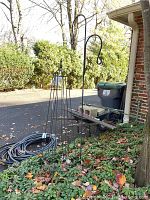 Photo showing black metal freestanding trellis and two-sided shepherd's crook plant holders outdoors near driveway.