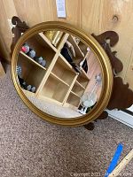 Gold-tone oval plastic frame mirror with reflective glass face shown leaning against a wood-paneled wall. The mirror is oval, about 30.5" x 24", with no visible damage.