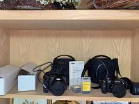 Overview on shelf: two white cardboard boxes, Nikon DSLR camera body with lens cap and strap, Nikon manual, yellow packaged Li-ion battery, Panasonic Lumix charger and two black fabric cases in background.