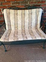 Front view of green metal loveseat with tufted beige striped cushions against brick background.