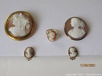 Group photo showing all five cameo jewelry pieces: two larger brooches or pendants, one small ring, and two small earrings laid out on white background