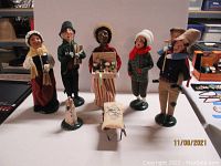 Full set showing five Byers' Choice Caroler Figurines, wood cart with sack of bird seed, and sitting dog figurine on a white surface.