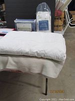 Beige quilted bedspread folded on a table with sheet sets and pillowcases in the background.