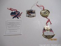 Four ornaments photographed on white background. Includes a flag-shaped ornament with blue ribbon dated 9/11, a White House Christmas Ornament with eagle and flags, a patriotic map-shaped ornament, and a circular gold ornament.