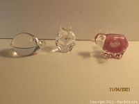 Front view of three glass figurines: a clear rounded pig on left, a clear frog or owl-shaped figure in center, and a pink pig on right, all on white surface with soft shadows.