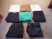 Eight pairs of folded women's pants in various colors (white, black, navy, tan, green) laid on floor