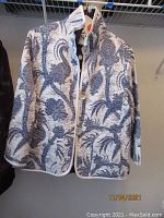 White and blue patterned coat with large floral/abstract design, quilted, hung on hanger.