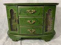 Front view of the green wooden jewelry box showing three gold-handled drawers and two side doors with gold mesh.
