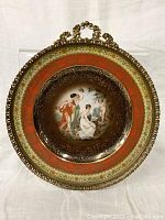 Full view of the plate showing central classical scene with three women and cherub, bordered by ornate gold and colored filigree and set in an elaborate metal frame.