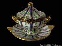 View of the two-piece hand painted Limoges porcelain pill box shaped like a tureen with lid and saucer showing floral decoration and vertical stripes with gold accents.