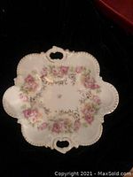 Front view of scalloped white porcelain plate decorated with pink roses and gold accents, showing two handles.