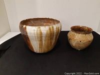 Two pottery pots displayed on black surface. Larger pot is mostly white with brown vertical glaze streaks; smaller pot is brown with horizontal white lines.