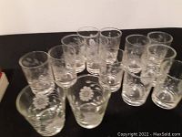 Twelve clear glasses arranged in a semi-circle showing the etched sunflower pattern on each glass side.
