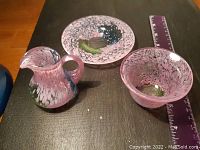 Three small art glass pieces: pitcher, bowl, and plate with mottled pink and other colors.