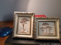Two framed decorative tiles showing relief tree designs, inside ornate gold frames and cream mats, set on a wooden surface.