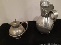 Side view of two metal jugs: one tall with ornate spiral handle, one shorter with hinged lid and spout
