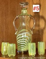 The full view of the figural art glass decanter with head stopper and four shot glasses arranged in front.