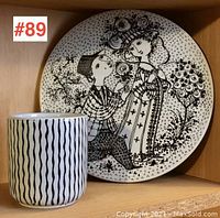 Two pieces of Nymolle pottery placed together on a wooden shelf, showing the cup with a black wavy line pattern and the black-and-white wall plaque with figures.