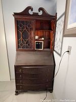 Front view of antique secretary desk showing closed hutch with decorative wooden lattice over glass, closed fold-down writing surface, and four drawers with round metal pulls; dark stained wood finish visible.