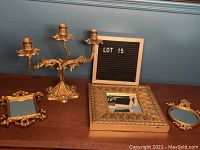 Photo showing one gold-colored metal three-arm candelabra along with three small decorative gold-colored mirrors on a wooden surface against a blue wall.