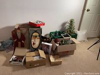 Overview photo showing various Christmas decorations, including a vintage wooden Santa figure, framed display, and several boxes with holiday decorations.