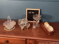 Full view of lot with various glass items on wooden surface: basket dish with handle, two large candy dishes, pair of glass doorknobs, and coasters in wicker holder.