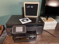 HP 6250 all-in-one printer/scanner/copier and instruction disk on top, set on a wooden table against a wall with a small display board showing lot number 92.