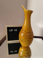 Front and side view of a tall yellow ceramic vase with brown stripes and a pointed, flared lip, standing on a mirror surface.