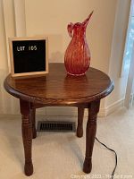Oak round end table in medium brown finish, showing wood grain and carved legs, with a pink and white glass vase on top and a letter board indicating lot number.