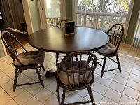 Full view of round oak table with 4 Windsor back chairs around it, all chairs with cushions on seats.