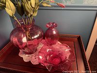 Trio of pink glass items including two vases and one ruffled candy dish arranged on wooden surface with lace doily.