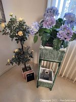 Wicker planter stand painted blue with faux purple and blue flowers next to pink ceramic planter with faux yellow roses.