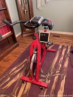 Full angled view of the red Soozier stationary exercise bike showing the frame, handlebars with LCD display, and pedals on a patterned rug indoors.