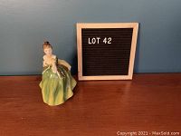 Full view of the Royal Doulton porcelain figurine 'Fleur' seated in a green dress next to a sign reading LOT 42