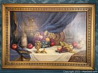 Full view of the framed still life print showing a table covered with a white cloth holding decorative metal vessels and various fruit including apples and grapes, with a blue draped background.