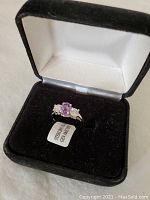 Amethyst sterling silver ring displayed inside black jewelry box with white interior lining. Ring shows oval purple amethyst center stone with two clear stones on either side. Tag attached labeled 'STERLING SILVER ON...'.