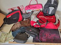 Photo showing assortment of bags including State Farm duffel, messenger bags, camera backpacks, and totes on bed surface