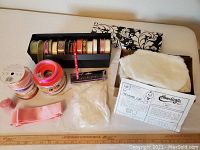Image showing assorted ribbons in different colors, some rolled on spools in a black storage box and some loose, alongside white stuffing and white faux fur fabric pieces.