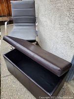 Open view of dark brown faux leather storage bench with hinged lid raised showing spacious internal storage compartment. Bench has stitched panel details on exterior surface.