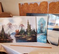 Four large paintings including two original landscape paintings by Miller shown on a table, view mostly front with varying perspectives