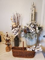 Photo showing rustic decor with a large winter faux floral arrangement with pinecones and plaid fabric ornaments in a metal container with rope handles, cotton plant arrangements in two vases, and an antique woven wicker basket with cotton stems.