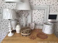 Photo showing white ceramic lamps, fabric box, wicker basket, white ornate frame, and pink glassware on wooden table.