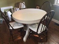 Photo showing round white painted hardwood dining table surrounded by four dark wood spindle back chairs with beige cushioned seats in a dining room with wooden floor.