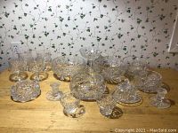 Full view of crystal glassware collection on wooden table against floral wallpapered wall. Includes bowls, stemmed glasses, covered dishes, and candle holders.