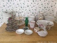 Wide view of assortment of decorative ceramics and glass items on wooden surface against floral wallpaper background.
