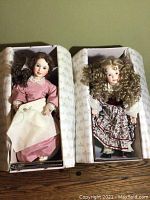Two vintage porcelain dolls in original boxes, one with brunette ringlets and pink dress, the other with curly blonde hair and floral dress.