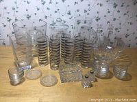 Wide angle photo showing various clear glassware items arranged on a wooden surface against a floral wallpaper background. Includes multiple cylindrical glasses stacked in groups, square containers, small bowls, and coasters.