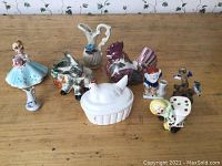 Group photo showing eight various decorative items including figurines, a small pitcher, and a lidded container shaped like a hen, arranged on a wooden surface.