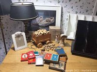 Photo showing assorted deck of cards, wooden block wagon, decorative figurines, lampshade, picture frames, and black display rack on wooden surface with floral wallpaper background.